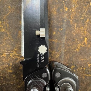 Peut inclure: Un outil multifonction pliable doté d'une lame noire avec un tranchant argenté. La lame porte l'inscription "USA" et "MagnaCut" et comprend un bouton de pouce blanc. Le manche est en métal gris foncé avec des vis visibles et une finition texturée.