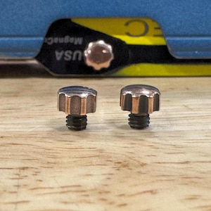 Könnte beinhalten: Zwei kleine Metall-Rändelschrauben mit kupferfarbenen, geriffelten, abgerundeten Köpfen und dunklen Gewindeschäften. Die Schrauben stehen aufrecht und zeigen ihre markante metallische Oberfläche sowie das strukturierte Design der Schraubenköpfe.