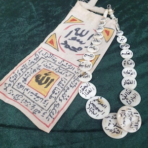 Op de afbeelding: Een gebroken witte religieuze amulet met Arabische schrift en een kralenketting van witte, cirkelvormige gebedskralen. De amulet heeft een rechthoekig stoffen zakje met tekst en geometrische patronen.