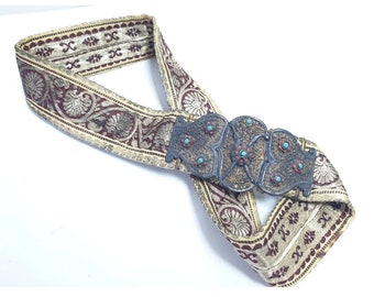 May include: A decorative belt with a silver buckle adorned with turquoise stones. The belt is made of a cream-colored fabric with a burgundy floral pattern.
