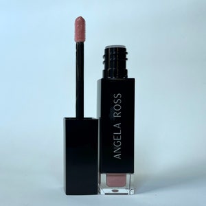 May include: A black rectangular tube of liquid lipstick with the brand name "ANGELA ROSS" in white lettering. The applicator is extended, showing a light pink lipstick colour. The background is white.