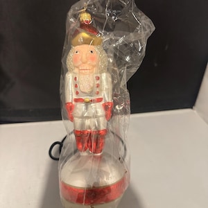 May include: A Christmas ornament shaped like a nutcracker soldier, encased in plastic. The ornament features a white uniform with red accents, a gold hat, and a stern expression. The base is white with a red band.