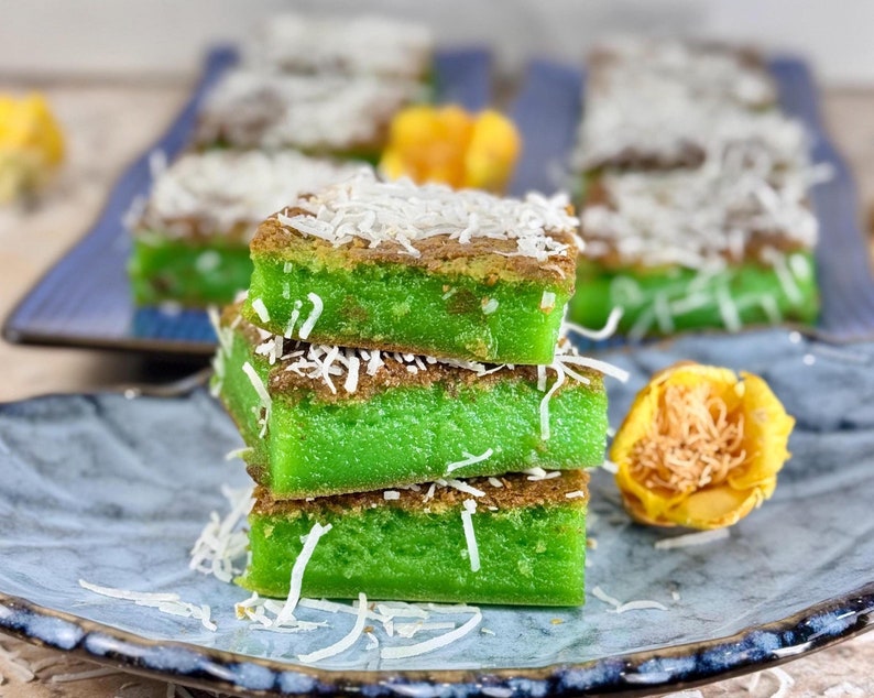 May include: A stack of three green and brown coconut cake squares on a blue plate. The cake is topped with shredded coconut.