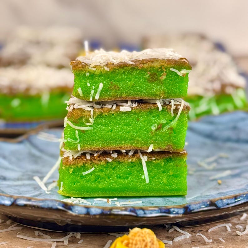 May include: A stack of three green and brown sponge cakes topped with shredded coconut. The cakes are on a blue and white patterned plate.
