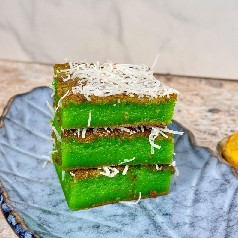 May include: Three layers of green pandan cake topped with shredded coconut on a blue and white plate.