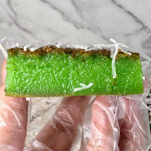 May include: A green and brown cake topped with shredded coconut. The cake is being held by a gloved hand.