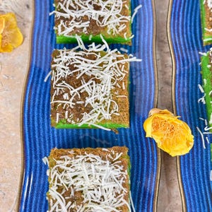 May include: Three squares of green cake topped with shredded coconut on a blue and white striped plate.