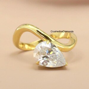 May include: A gold-colored ring with a pear-shaped, clear gemstone. The ring's band has a unique, flowing design. The ring is set against a soft, neutral background, with the text "CrushedIceDiamonds" visible.