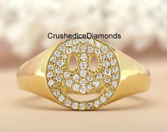 Puede incluir: Un anillo de oro con un diseño de carita sonriente. La carita sonriente está hecha de pequeñas piedras brillantes. El anillo tiene una banda lisa y pulida y una cara redondeada. El texto "CrushedIceDiamonds" es visible en la imagen.
