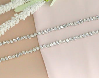 May include: Two diamond necklaces with various cuts, including pear and emerald shapes, are displayed on a pink surface. The necklaces are silver-toned and feature multiple clear gemstones. White floral accents are visible in the background. The text "CrushedIceDiamonds" is in the lower left corner.