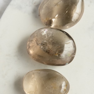 Peut inclure: Trois pierres lisses, de couleur brun clair, de forme ovale et polies. Les pierres sont disposées verticalement, la plus grande en haut et la plus petite en bas.