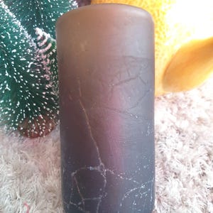 May include: A dark grey pillar candle with a cracked, textured surface. The candle is cylindrical and stands upright. A small, green Christmas tree and a yellow teapot are in the background.