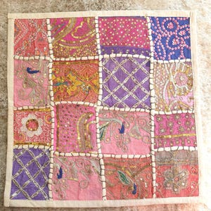 Op de afbeelding: Een vierkante patchworktextiel met roze, paarse en gouden stoffen. Elk vierkant is versierd met pailletten en borduursels. De textiel is omrand met een crèmekleurige stof.