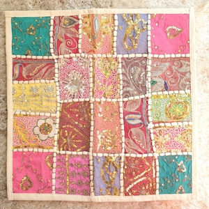 Peut inclure: Textile carré en patchwork avec une bordure crème. Le motif présente une grille de carrés colorés dans les tons de rose, turquoise, or et violet, ornée de sequins et de broderies. Il s'agit probablement d'un coussin décoratif.