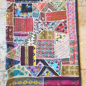 Puede incluir: Un textil de patchwork colorido con un borde negro. El textil está compuesto por varias piezas de tela en una variedad de colores y patrones, incluyendo diseños florales, geométricos y abstractos. El textil es de forma rectangular.