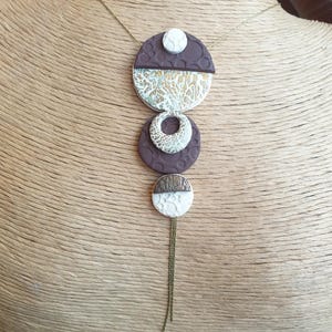 Peut inclure: Un collier unique avec des formes géométriques marron, blanches et dorées. Le pendentif est composé de plusieurs couches et est suspendu à une longue chaîne avec une petite tresse. Le collier est présenté sur un mannequin.