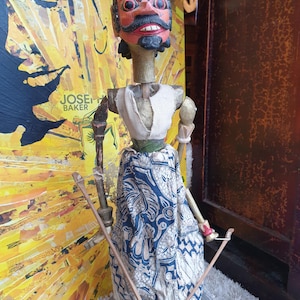 Peut inclure: Une marionnette en bois avec un visage peint et une couronne. La marionnette porte une jupe à motifs et tient deux bâtons en bois. La marionnette est debout sur une base ronde.