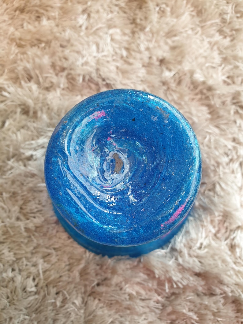 May include: A round, blue decorative object with a swirling pattern of white, pink, and blue paint. The object has a glossy finish and sits on a fluffy, light-colored surface. The object's design is reminiscent of a galaxy.
