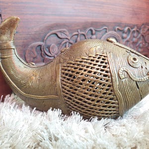Peut inclure: Une sculpture de poisson dorée détaillée avec une surface texturée et un design ajouré. Le poisson a une queue incurvée et des nageoires détaillées. La sculpture est posée sur une surface blanche et moelleuse, avec un fond en bois visible.