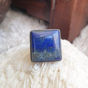 Peut inclure: Une bague carrée en lapis-lazuli bleu foncé avec des mouchetures dorées. La pierre précieuse est sertie dans un métal de couleur argent. La bague est présentée sur une surface blanche et texturée, peut-être du corail.