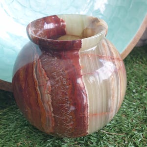 Peut inclure: Un vase sphérique fabriqué en pierre rubanée polie avec une finition lisse et brillante. La surface présente une palette de couleurs naturelles composée de couches brun-rouge profond, crème et vert pâle translucide avec des veines minérales.