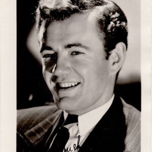 May include: Black and white photo of a man in a suit smiling and looking at the camera. He is wearing a white shirt and a dark tie. The photo is signed "much" in the bottom left corner.
