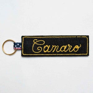 May include: A rectangular black felt keychain featuring the word Camaro embroidered in yellow cursive script. The keychain has a yellow stitched border and is attached to a gold-toned metal ring by a fabric tab patterned with white stars.