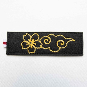 Op de afbeelding: Een zwart vilten rechthoek met gouden borduursels van bloemen- en wolkenmotieven. Een klein rood-wit gestreept lint is aan de linkerkant bevestigd. Het borduurwerk is gedetailleerd en het ontwerp is gecentreerd.