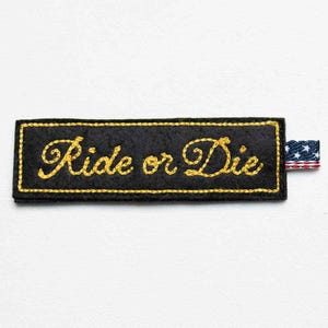 May include: A rectangular black fabric patch featuring the embroidered yellow text Ride or Die in a cursive script. The patch is outlined with a yellow stitched border and includes a small tab on the right side displaying a red, white, and blue flag pattern.