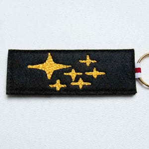May include: A rectangular black felt keychain featuring embroidered gold stars of varying sizes. The design includes one large star and five smaller four-pointed stars. A small white and red striped fabric tab is attached to the side with a metal ring.