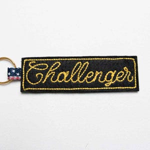 May include: A rectangular black fabric keyring featuring the word Challenger embroidered in yellow cursive script. The keyring is attached to a gold-toned metal ring by a small fabric tab patterned with the American flag stars and stripes.