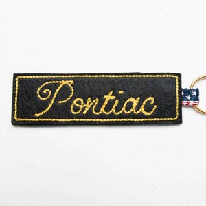 May include: A rectangular black felt keychain featuring the word Pontiac embroidered in yellow script. The border is stitched in yellow thread, and a small fabric tab with a red, white, and blue star pattern is attached to a gold metal ring.