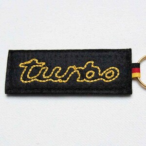 May include: A rectangular black felt keychain featuring the word Turbo embroidered in yellow thread. The keychain includes a small fabric tab with black, red, and yellow horizontal stripes attached to a gold-toned metal split ring.