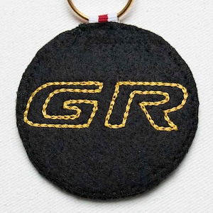 May include: A circular black felt keychain featuring the letters GR embroidered in yellow thread. The edges of the felt are stitched with black thread, and a gold-toned metal ring is attached to the top with a small red and white fabric tab.