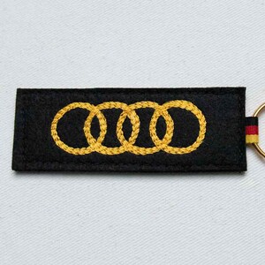 May include: A rectangular black felt keychain featuring four interlocking yellow embroidered rings. On the right side, a small fabric tag displays horizontal stripes in black, red, and yellow, attached to a gold-toned metal ring.