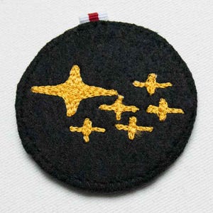 May include: A circular black felt patch featuring embroidered yellow stars of varying sizes. The design includes one large four-pointed star and five smaller four-pointed stars arranged in a cluster. A small white and red fabric tag is attached at the top.