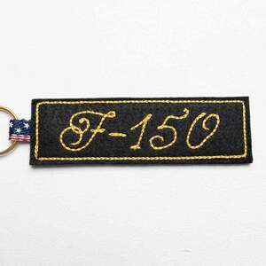 May include: A rectangular black felt keychain featuring the text F-150 embroidered in yellow cursive thread. The border is stitched in yellow, and a small fabric tab with a red, white, and blue American flag pattern is attached to a gold metal ring.
