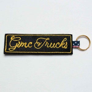 May include: A rectangular black fabric keychain featuring the yellow embroidered text "Gmc Trucks" in a cursive script. The keychain is bordered by yellow stitching and includes a small fabric tab with a stars and stripes pattern attached to a metal ring.