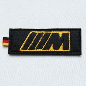 May include: A rectangular black felt patch featuring an embroidered yellow letter M with three diagonal stripes. A small fabric tab with horizontal black, red, and yellow stripes is attached to the left side of the patch, which has stitched edges.