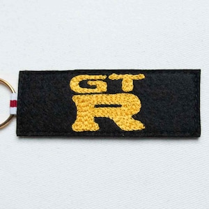 May include: A rectangular black fabric keychain featuring the yellow embroidered text GT-R in the center. The keychain is attached to a circular metal ring with a small white and red striped fabric tab connecting the two components together.