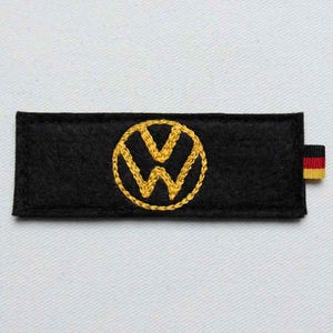 May include: A rectangular black felt patch featuring a centered, circular yellow embroidered Volkswagen logo. The patch has a small fabric tag on the right side with horizontal black, red, and yellow stripes, representing the German national flag.