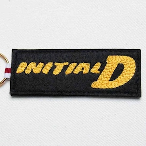 Peut inclure: Porte-clés rectangulaire noir avec le texte "INITIAL D" brodé en or. Le porte-clés a un anneau doré et un petit accent rayé blanc, rouge et blanc. Le fond est une surface blanche unie.