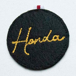 May include: A circular black felt patch featuring the word Honda embroidered in yellow cursive script. The patch has a small red and white fabric tab at the top center, and the edges are finished with black stitching around the entire circumference.