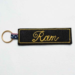 May include: A rectangular black felt keychain featuring the name Ram embroidered in yellow cursive script. The keychain is attached to a gold-toned metal ring by a small fabric tab patterned with white stars on a blue background and red stripes.