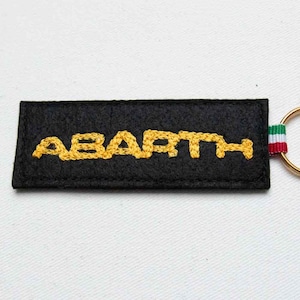 May include: A rectangular black felt keychain featuring the word ABARTH embroidered in yellow thread. The keychain includes a small fabric loop with green, white, and red horizontal stripes attached to a gold-toned metal split ring.