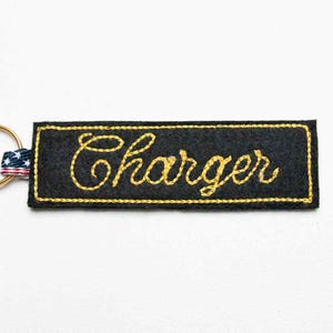 May include: A rectangular black felt keyring featuring the word Charger embroidered in yellow cursive script. The edges are stitched with yellow thread, and a metal ring is attached to the left side with a small American flag patterned fabric tab.