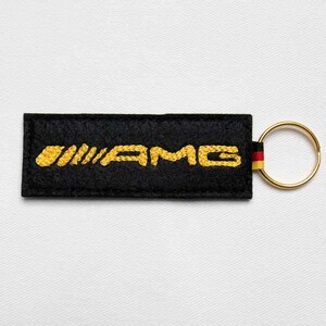 May include: A rectangular black felt keychain featuring the yellow embroidered text AMG. The keychain includes a gold-toned metal split ring attached by a small fabric tab displaying the black, red, and yellow colors of the German flag.