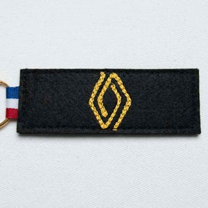 May include: A rectangular black felt keychain featuring a yellow embroidered diamond-shaped geometric pattern in the center. On the left side, a small fabric loop displays horizontal stripes in blue, white, and red, attached to a gold metal ring.