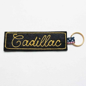 May include: A rectangular black felt keychain featuring the word Cadillac embroidered in yellow cursive script. The keychain includes a small fabric tab with a red, white, and blue star-spangled pattern attached to a circular gold-tone metal ring.