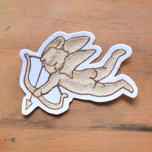 Peut inclure: Écusson brodé représentant Cupidon, une figure ailée avec un arc et une flèche. Le patch est doré avec une bordure blanche, représentant Cupidon en vol. Idéal pour personnaliser les vêtements ou les accessoires, cet écusson est un élément décoratif charmant.